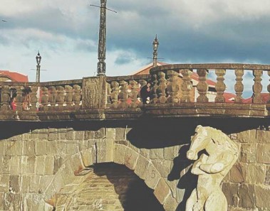 A tikbalang statue stands with arms crossed near the lower arch of a medium sized stone bridge.