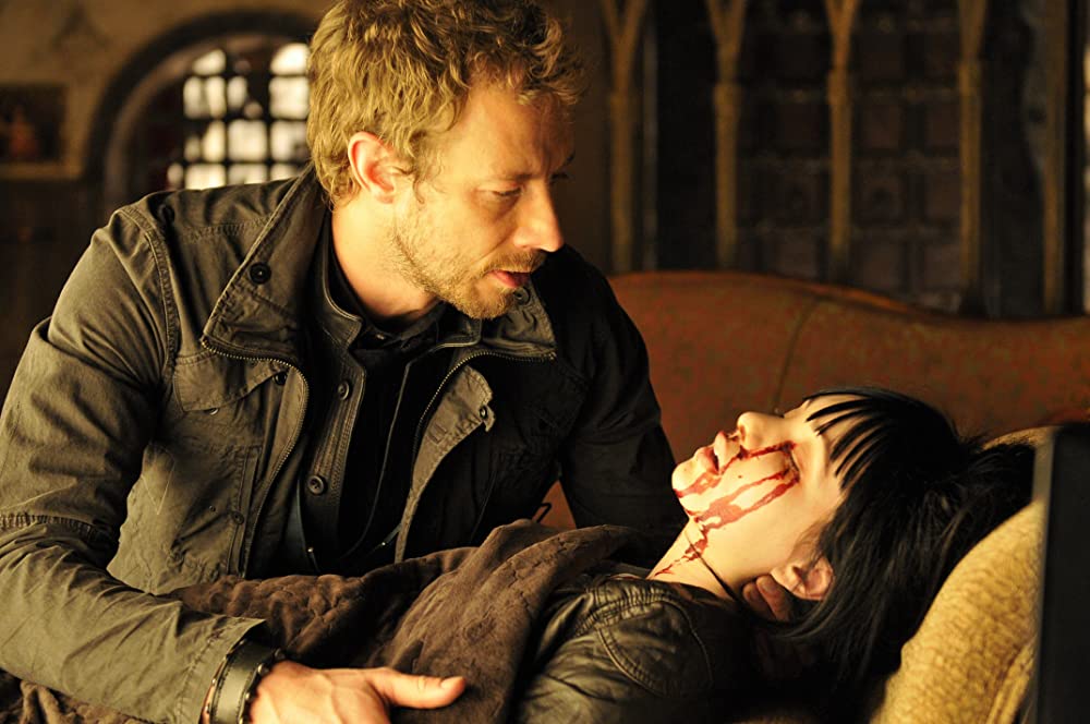 Still from Lost Girl episode. A girl lay ill on a couch, bleeding from the eyes, as a man sits beside her in a caring manner. 