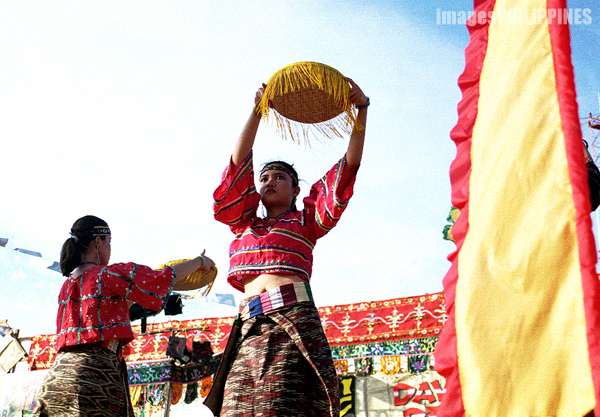 A T'boli girl in traditional attire holds a basket over her head while performing a ritual.