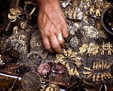 A hand reaches into a pile of various metal medallions of different shapes, symbols, and sizes, mixing indigenous, Catholic, and other imagery.