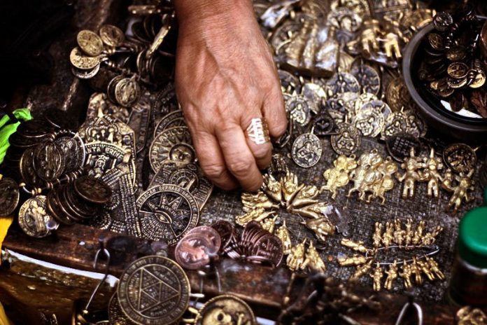 A hand reaches into a pile of various metal medallions of different shapes, symbols, and sizes, mixing indigenous, Catholic, and other imagery.