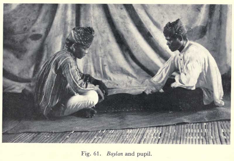 Two men sit cross legged on the floor. They wear tradtitional bukidnon clothing, with loose fitted shirts and a wrap on their heads.