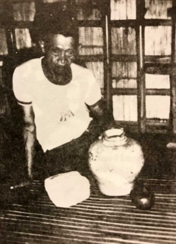 A man sits on a bamboo floor with ritual jars in front of him.