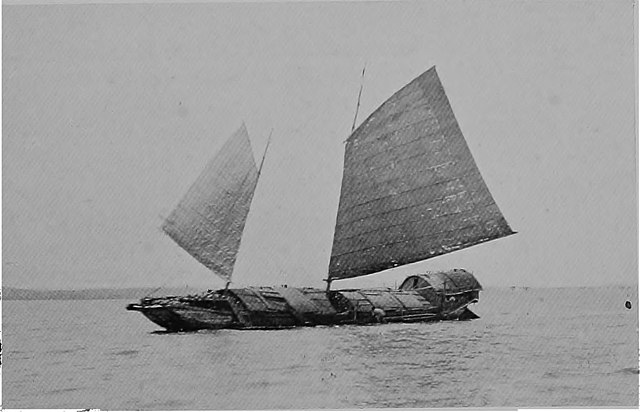 A wooden boat with a flat front and two sails is moving across the ocean.