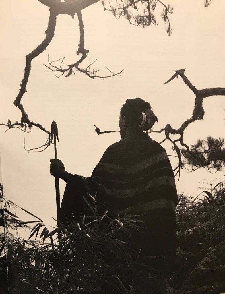 A traditionally dressed Bontoc man sits under a tree, smoking his pipe, overlooking the moutains of the Philippine Cordillera.