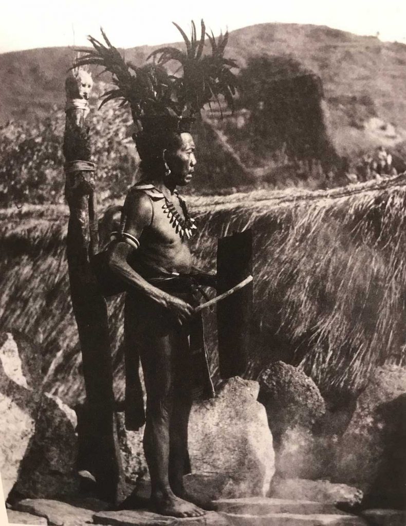 A traditionally dressed bontoc man stands near a totem, calling out for the warriors to come to the village center.