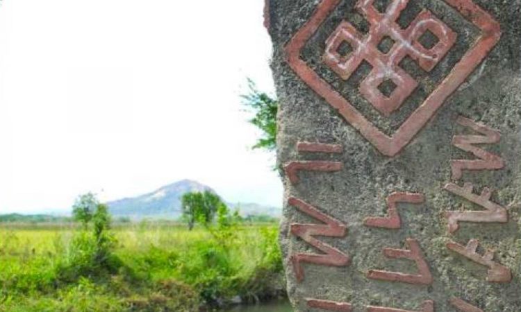 A stone marker shows traditional buhid syllabary with a tribal symbol. The village and mountains are seen in the background.