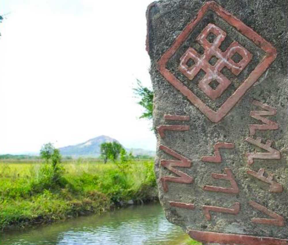 A stone marker shows traditional buhid syllabary with a tribal symbol. The village and mountains are seen in the background.